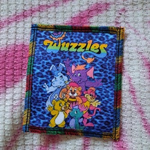 Könnte beinhalten: Ein rechteckiger Aufnäher mit dem Wort "Wuzzles" in Regenbogenbuchstaben und Zeichentrickfiguren auf blauem Leopardenmuster. Der Aufnäher hat einen Regenbogenrand.