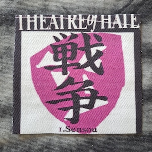May include: A square patch with the text "THEATRE of HATE" in white capital letters above a pink shield shape containing black Japanese characters. The text "I. Sensou" is below the shield.