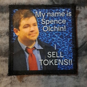 May include: A rectangular patch featuring a portrait of a man in a suit and tie. Text above the portrait reads "My name is Spence Olchin!" and below it says "I SELL TOKENS!!" The background is blue with a leopard print.