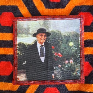 May include: Square patch with a printed image of a man in a suit and hat, standing in front of a garden with red roses. The patch has a red stitched border and is set against a patterned orange and black background.