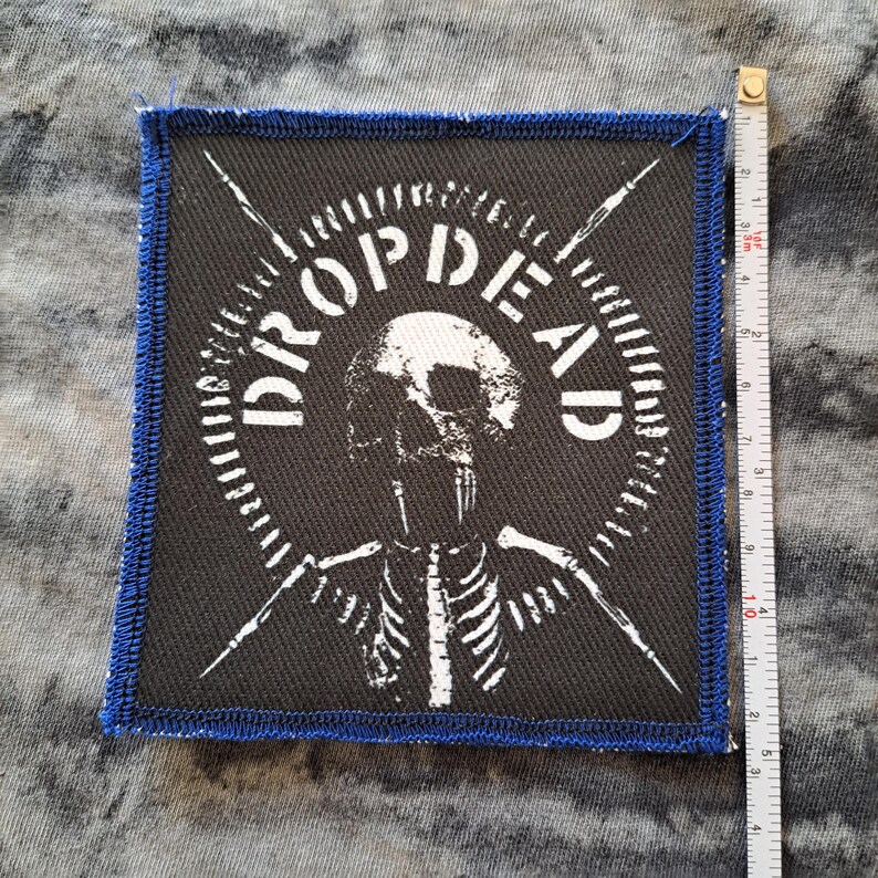 May include: Square black patch with a blue stitched border. The patch features the white text "DROPDEAD" in a circular design around a skull and skeleton illustration. Two white arrows are crossed behind the skull. The patch measures approximately 4 inches by 4 inches.