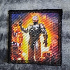 Puede incluir: Parche cuadrado con Robocop sosteniendo un arma, sobre un fondo vibrante y ardiente. El parche, con borde negro, está diseñado para aplicarse en ropa o accesorios.