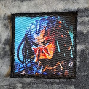 May include: Square patch featuring a detailed image of the Predator alien from the movie. The patch has a black border and a textured background. The Predator is depicted with blue, orange, and black colors.