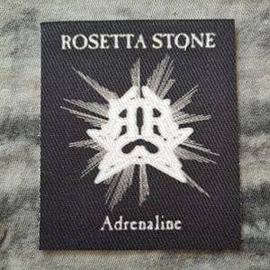 May include: A black rectangular label with the text "ROSETTA STONE" at the top and "Adrenaline" at the bottom. A white, stylized graphic is in the center. The label is on a textured, gray surface.