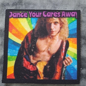 May include: Square patch with a black border featuring a portrait of a person with long blonde hair and the text "Dance Your Cares Away" in purple. The background is a rainbow design.