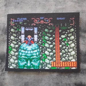 May include: A rectangular patch featuring a pixelated video game scene. The scene includes a character atop a green stone head, a red ladder, and text that reads "PLAYER" and "ENEMY". The background is a stone wall.