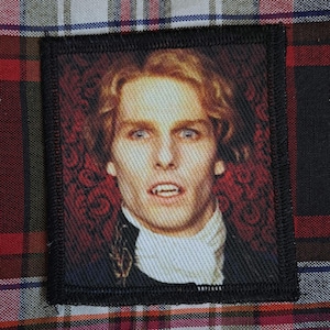 May include: A square fabric patch featuring a printed portrait of a person with wavy light brown hair, pale skin, and visible fangs. The individual wears a white cravat and a dark coat against a dark red patterned background with a black stitched border.