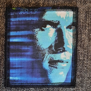 May include: A square fabric patch with a black stitched border featuring a blue and black high-contrast portrait of a person. The left side displays horizontal blue lines and the text "ON TO WIN AT ANY COST" and "WHO WILL WIN THIS".