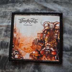 Puede incluir: Parche cuadrado con borde negro que presenta una ilustración detallada. La obra de arte representa una figura de calavera con cuernos con otras calaveras y una fortaleza en tonos naranja, marrón y blanco. El texto "Dimmu Borgir" y "bledsvept" está en la parte superior.