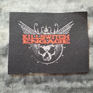 May include: A rectangular black patch with the words "KILLSWITCH ENGAGE" in orange letters. The patch features a white skull with wings and a crown design. The patch is likely for a band or music merchandise.