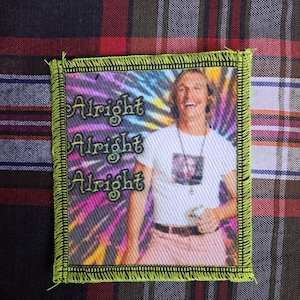 May include: A square patch with a neon green border and the words "Alright Alright Alright" in a stylized font. The patch features a man smiling against a tie-dye background. He is wearing a white t-shirt and pink pants.