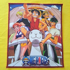 May include: A rectangular patch featuring a colorful illustration of characters from the anime series One Piece. The design includes a ship's helm, a sheep-shaped figurehead, and the series' logo with a skull and crossbones. The patch has a red border.