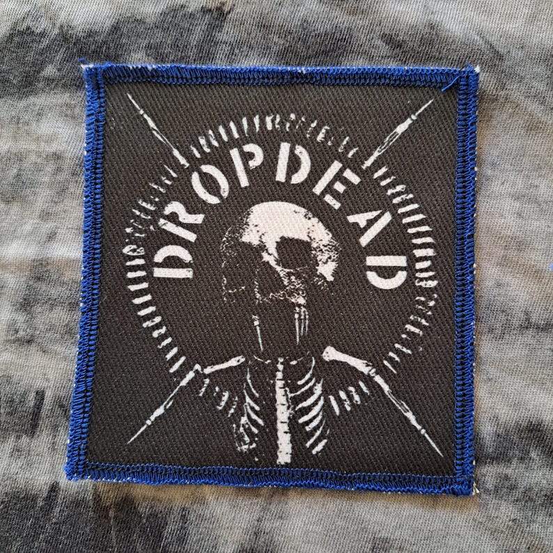 May include: Square black patch with a blue stitched border. The patch features a white skull and skeleton design with the words "DROPDEAD" in a circular pattern. Two white crossed arrows are also present.