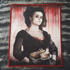 May include: A rectangular patch featuring a portrait of a woman in a dark dress holding a plate of food. The background has red streaks. The patch has a red border.