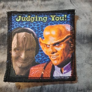May include: Square patch with a black border featuring two characters from a science fiction series. The patch has the text "Judging You!" in green. One character is a light-skinned humanoid, and the other is orange-skinned with pointed ears.