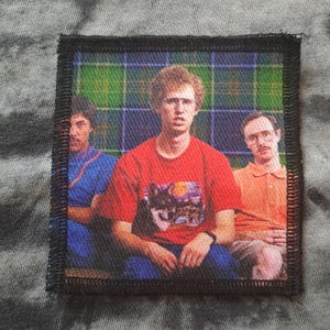 May include: Square patch featuring a group of three people. The central figure wears a red t-shirt with a graphic, while the others are in blue and orange. The background has a plaid pattern. The patch is bordered with black stitching.