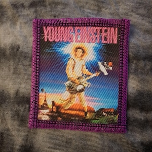 May include: A rectangular patch with a purple border and the text "YOUNG EINSTEIN". The image depicts a person playing a guitar with a bright light behind them. The background shows a landscape with buildings and a sunset.