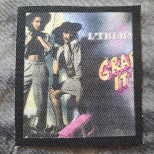 May include: A rectangular fabric patch with a black border. The patch features a colorful image of two people wearing hats and stylish clothing. The text "L'TRIM" and "GRAB IT!" are visible on the patch.