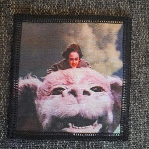 May include: A square fabric patch with a black stitched border featuring a printed image of a person with dark hair riding on the back of a large, light-colored, furry creature with a wide, smiling mouth against a cloudy, light-colored sky.