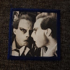 May include: A square fabric patch featuring a black and white photographic print of two individuals facing each other in profile. The patch has a dark blue stitched border that frames the image, creating a distinct edge around the central graphic.