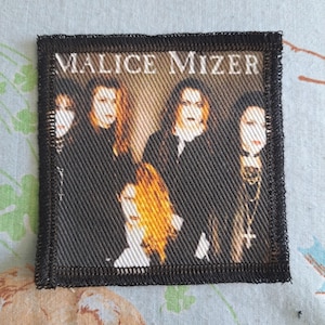 May include: Black and white patch featuring the band Malice Mizer. The patch has a black border and displays a group photo of the band members. The band's name is printed in white at the top.