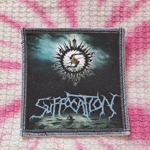 May include: A square, black patch with a detailed design. The patch features a central, spiky sun-like emblem above the word "SUFFOCATION" in stylized, light blue lettering. The edges are finished with a stitched border.