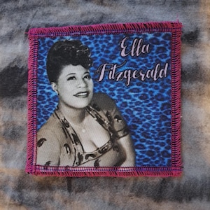 Puede incluir: Un parche cuadrado con una imagen en blanco y negro de Ella Fitzgerald, con el nombre "Ella Fitzgerald" en escritura cursiva. El fondo es azul con un diseño de estampado de leopardo, y el parche tiene un borde cosido rosa.
