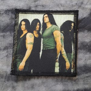 May include: A rectangular patch featuring a band of four men with long dark hair. The men are wearing black and green tops. The patch has a black border and is set against a gray, tie-dye background.