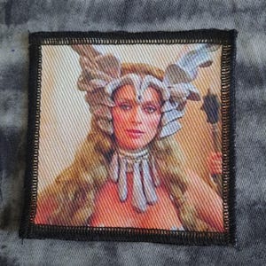 May include: Square patch with a portrait of a woman with long blonde hair and ornate headwear. The image has a vintage feel, with a black border and a fabric texture. The woman wears a necklace and has a serious expression. The patch is square.