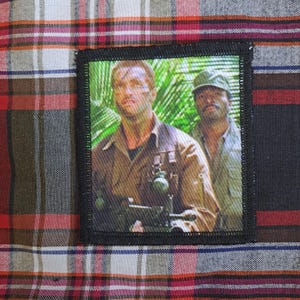 May include: A rectangular patch featuring a scene from a movie, with two men in military attire against a jungle backdrop. The patch is bordered in black and is set against a plaid fabric background with red, white, and black colors.
