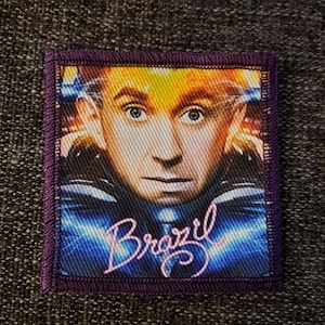 Op de afbeelding: Een vierkante stoffen patch met een close-up portret van een persoon met licht haar en blauwe ogen, tegen een donkerblauwe achtergrond met lichtstrepen. Het woord Brazil staat onderaan in een lichtgekleurd cursief lettertype.