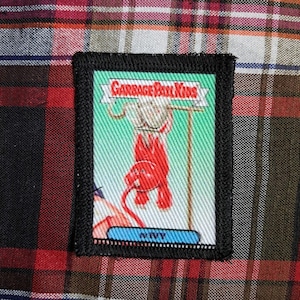 Può includere: Una toppa rettangolare con bordo nero con un'illustrazione di Garbage Pail Kids. L'immagine mostra una figura rossa che tiene in mano un cartello con la scritta "Garbage Pail Kids". La parte inferiore della toppa riporta la parola "NIVY" in un riquadro blu.