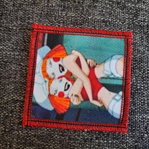 May include: A square fabric patch with a red stitched border featuring a printed illustration of two figures with orange hair, pale skin, and red cheek markings, wearing red clothing while embracing against a teal and white background.