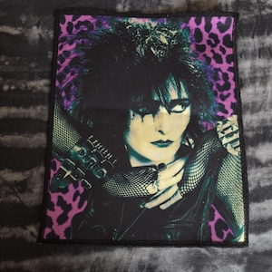 May include: A rectangular patch featuring a portrait of a person with dark hair and makeup, set against a purple and black leopard print background. The person is wearing a black jacket and mesh gloves. The patch has a black border.