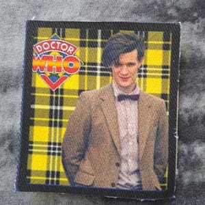 May include: A white ceramic mug with a black handle and the text "But First, Coffee" in a bold, black font. The mug is set against a blurred background, suggesting a cozy setting.