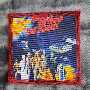 Könnte beinhalten: Quadratischer Aufnäher mit rotem Rand. Der Aufnäher zeigt den Text "Battle Beyond The Stars" über einer Weltraumszene mit Figuren und Raumschiffen. Der Hintergrund ist blau, gelb und orange.