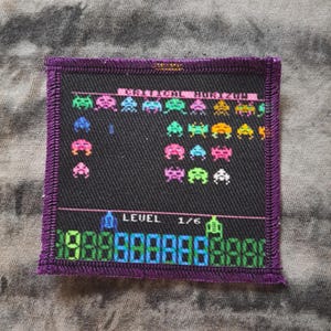 May include: Square patch with a retro video game design. The patch features colorful pixelated alien figures and game elements on a black background, framed by a purple border. The text "LEVEL 1/6" is visible.