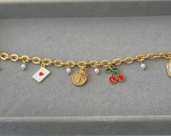 May include: Gold-toned charm bracelet featuring a series of decorative charms. Charms include a cowboy boot, a playing card with a heart, a cross, cherries, and a floral design. Small white pearls are interspersed along the chain.