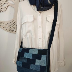 Peut inclure: Sac messager en patchwork de denim avec fermeture à rabat et bouton en bois avec un motif d'arbre. Le sac a une bandoulière bleu foncé et est présenté avec une chemise blanche et une écharpe bleu marine.