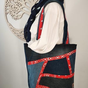 Peut inclure: Un sac fourre-tout en denim avec un design patchwork. Le sac présente différentes nuances de denim bleu, rehaussées de tissu paisley rouge. Les sangles et les bords sont garnis de tissu paisley rouge. Le sac est porté par une personne vêtue d'une chemise blanche et d'une écharpe bleu foncé.