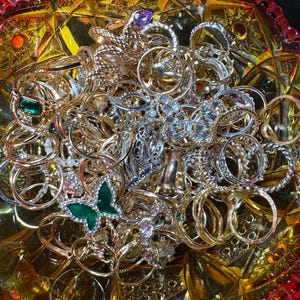 K&ouml;nnte beinhalten: Eine Sammlung verschiedener Ringe in Gold- und Silbert&ouml;nen, einige mit Edelsteinen verziert. Die Ringe werden in einer dekorativen, bernsteinfarbenen Glasschale pr&auml;sentiert. Mehrere Ringe haben aufwendige Designs, darunter ein gr&uuml;ner Schmetterlingsring.