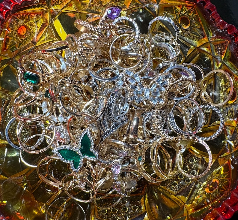 K&ouml;nnte beinhalten: Eine Sammlung verschiedener Ringe in Gold- und Silbert&ouml;nen, einige mit Edelsteinen verziert. Die Ringe werden in einer dekorativen, bernsteinfarbenen Glasschale pr&auml;sentiert. Mehrere Ringe haben aufwendige Designs, darunter ein gr&uuml;ner Schmetterlingsring.