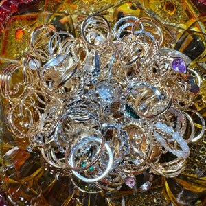 K&ouml;nnte beinhalten: Eine Sammlung goldener und silberner Ringe in verschiedenen Designs, darunter B&auml;nder, verzierte Stile und Ringe mit Edelsteinen. Die Ringe sind in einer dekorativen, strukturierten Glasschale angeordnet und bilden eine funkelnde Schmuckausstellung.