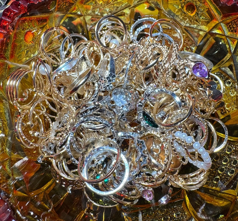 K&ouml;nnte beinhalten: Eine Sammlung goldener und silberner Ringe in verschiedenen Designs, darunter B&auml;nder, verzierte Stile und Ringe mit Edelsteinen. Die Ringe sind in einer dekorativen, strukturierten Glasschale angeordnet und bilden eine funkelnde Schmuckausstellung.