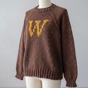 Peut inclure: Un pull en tricot marron avec un "W" jaune sur le devant. Le pull a un col rond, des manches longues et des poignets et un ourlet côtelés. Le tissu présente des mouchetures de couleur.