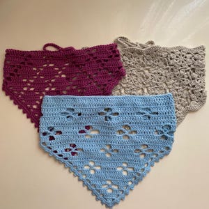Pode incluir: Três bandanas de crochê feitas à mão em tons de roxo, azul claro e bege. Cada bandana apresenta um design vazado com uma borda ondulada. As bandanas são em forma triangular e parecem ser feitas de fio macio.