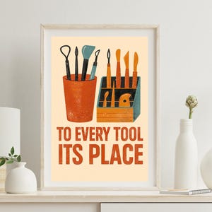 May include: Framed art print featuring pottery tools in a terracotta-colored cup and a wooden tool holder. The print has the text "TO EVERY TOOL ITS PLACE" in orange and white. The artwork is displayed on a white shelf.