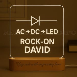 May include: A personalized LED night light with a wooden base. The clear acrylic panel displays the text "AC -> DC -> LED ROCK-ON DAVID" and an electrical symbol. The base is engraved with "Upgrade with engraving here".