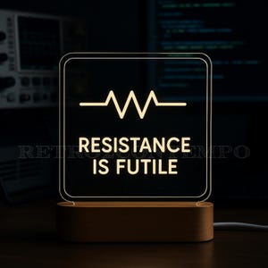 Peut inclure: Un panneau lumineux à LED avec le texte "RESISTANCE IS FUTILE" et un symbole de forme d'onde. Le panneau carré aux coins arrondis repose sur une base en bois. La lumière est de couleur blanc chaud.