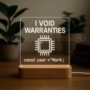 Peut inclure: Lampe de chevet LED rectangulaire avec une base en bois. Elle affiche le texte "I VOID WARRANTIES" au-dessus d'un graphique de puce informatique et le code "const user = 'Mark;'" en dessous. La lumière est allumée.